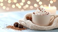 Hot chocolate topped with whipped cream and cocoa powder in white mug wrapped in knitted scarf, with spices and festive bokeh lights in background.