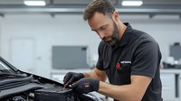 Caucasian male auto mechanic with beard wearing black polo shirt and protective gloves performing maintenance check under car hood in modern service garage.