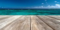 Wooden deck or pier overlooking turquoise ocean waters with blue sky and white clouds, selective focus on weathered planks in foreground for summer vacation concept.