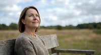 Middle-aged Caucasian woman in beige sweater sitting on wooden bench outdoors, looking thoughtfully into distance with peaceful expression against rural landscape.