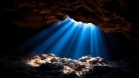 Dramatic blue light rays pierce through cave opening, illuminating rocky formations below against dark cavern background. Natural phenomenon creates mystical atmosphere.