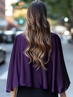 Young woman with long wavy ombre hair wearing purple cape-style top viewed from behind against blurred urban background with bokeh lights.