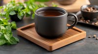 Black ceramic cup with hot coffee on wooden bamboo coaster, fresh mint leaves and roasted coffee beans in dark moody setting, selective focus composition.