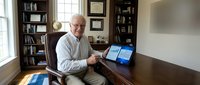 Senior Caucasian businessman working on laptop computer in home office library with wooden desk and bookshelves during remote work session.