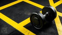 Single black dumbbell weight on dark gym floor with bright yellow line markings. Fitness equipment for strength training, weightlifting workouts, and exercise routines.
