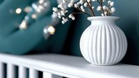 White ceramic vase with delicate branches and small white flowers on modern shelf against teal wall with warm bokeh lights creating cozy home decor atmosphere.