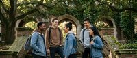 Diverse group of young college students having friendly conversation outdoors on university campus with historic stone architecture and green trees in background.