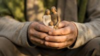 Senior man cupping hands protectively around wooden family figurines outdoors, symbolizing care, protection and family values in warm natural light.