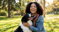 Happy young African American woman with curly hair embracing her black and white dog in sunny autumn park with golden light and fallen leaves.