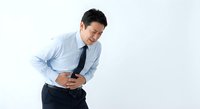 Asian businessman in blue shirt experiencing severe stomach pain holding abdomen with both hands against white background for medical concepts.