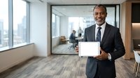 Mature African American male professional in formal business attire holding blank tablet in modern office with city skyline view and colleagues in background.