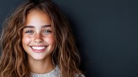 Young woman with wavy brown hair and bright genuine smile against dark background, radiating happiness and confidence in casual portrait style.