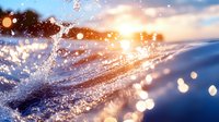 Ocean wave splashing at sunset with golden light reflecting off water droplets, creating sparkling bokeh effect against dramatic sky.