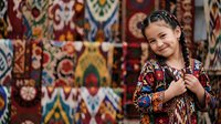 Young girl with dark hair in braids, wearing a colorful patterned traditional dress, smiling at camera against ornate woven textile backdrop. Cultural heritage portrait.