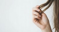 Woman examining damaged split ends of brown hair strand with fingers against neutral background for haircare and beauty treatment concepts.
