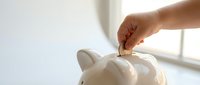 Hand inserting coin into white ceramic piggy bank for savings and financial planning concept on bright windowsill background.