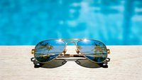 Aviator sunglasses with gold metal frames resting on wooden deck beside sparkling swimming pool, summer vacation and leisure concept.
