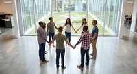 Diverse group of people holding hands in circle formation inside modern building with large windows for team building and unity concepts.