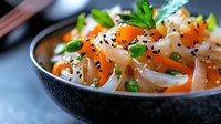 Fresh Asian noodle salad with carrots, green peas, sesame seeds, and herbs in a dark ceramic bowl, close-up food photography.