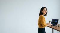 Young Asian woman in mustard yellow cardigan working at standing desk with laptop and coffee cup, looking upward thoughtfully in modern office workspace.