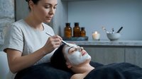 Professional esthetician applying facial mask treatment to relaxed female client in modern spa salon with skincare products and peaceful atmosphere.