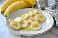 Fresh sliced bananas on white ceramic plate with honey drizzle, whole yellow bananas and striped kitchen towel in background on marble surface.