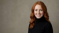 Professional redhead woman in black turtleneck smiling confidently against neutral background for business and corporate marketing materials.