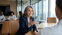 Mature businesswoman with gray hair in navy suit gesturing during meeting in modern conference room with colleagues in background.