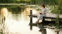 Young woman in white dress sitting on wooden dock reading book by peaceful lake at sunset surrounded by reeds and lily pads for relaxation and mindfulness.