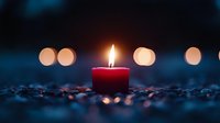 Red burning candle with soft glowing flame against dark background with blurred bokeh lights, romantic atmosphere on scattered coffee beans surface.