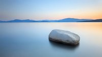 Serene lake at sunset with smooth stone in calm water, mountains silhouetted against pastel sky, peaceful minimalist landscape for meditation and wellness.