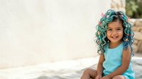 Young girl with teal curly hair wearing light blue sleeveless top, smiling outdoors in sunny setting, suitable for children's fashion, lifestyle, and diversity stock imagery.