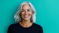 Confident mature woman with silver gray hair and natural smile wearing black top against turquoise background, representing aging gracefully and vitality in senior years.
