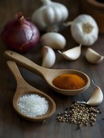 Fresh garlic cloves, red onion, wooden spoons with sea salt and turmeric powder, scattered coriander seeds on dark wooden surface in moody natural lighting.
