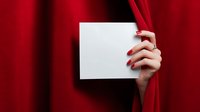 Female hand with red manicure holding blank white card against rich red velvet curtain background, creating dramatic contrast for announcement or invitation.