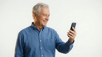 Senior man with gray hair and Caucasian ethnicity wearing blue denim shirt, holding and viewing smartphone against white background. Suitable for technology, communication, and lifestyle content.