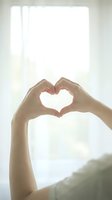 Young woman forming a heart shape with her hands against a bright window, soft light, love and care concept for Valentine or wellness.