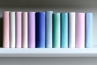 Row of hardcover books in pastel colors arranged on white shelf, spines showing gradient from pink through purple and blue to mint green, creating minimalist aesthetic.