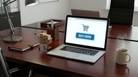Laptop on wooden desk displaying online shopping cart with buy now button, coffee cup and stationery in modern office interior.