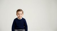 Young boy, approximately 5-7 years old, Caucasian ethnicity, wearing dark navy ribbed sweater, smiling at camera against neutral white background, studio portrait style.