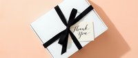 White gift box with elegant black ribbon bow and thank you card on pastel peach background, top view for holiday, wedding, or corporate gifting.