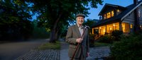 Senior businessman in elegant suit standing confidently on cobblestone driveway at twilight with luxury home glowing warmly in background.