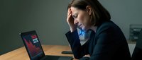 Stressed businesswoman holding head in hands while working on laptop in modern office, experiencing work pressure and mental fatigue during busy workday.