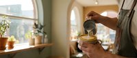 Hands pouring hot water into teacup in warm sunlit kitchen with window light, morning beverage preparation for tea time or breakfast routine.