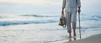 Senior man in light blue linen clothing walking along sandy beach shore with walking stick and bag, ocean waves in background, peaceful coastal lifestyle scene.