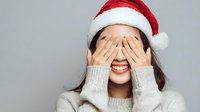 Young Asian woman in Santa hat covering eyes with hands while smiling, wearing gray sweater against neutral background.