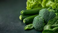 Fresh green vegetables including broccoli, zucchini, lettuce and spinach leaves arranged on dark textured background with copy space. Healthy eating concept.