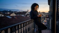 Young woman with curly dark hair and medium skin tone standing on urban balcony at dusk, wearing black jacket, gazing at cityscape with historic rooftops and distant mountains, suitable for lifestyle, travel, and urban living concepts.