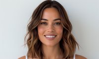 Young woman with wavy brown hair and highlights smiling confidently against white background, showcasing natural makeup and healthy skin.