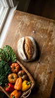 Rustic sourdough bread loaf with fresh organic vegetables in wicker basket on weathered wooden table by window for healthy farm-to-table cooking concept.